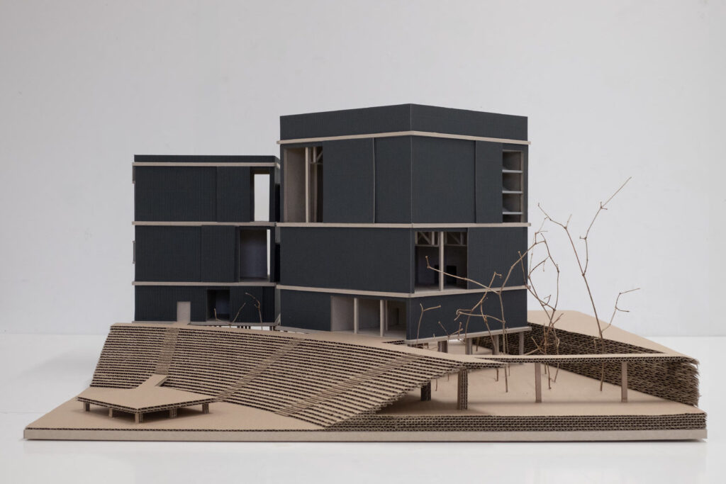 Architekturmodell eines modernen, kantigen Gebäudes mit dunkler Fassade auf einer geneigten Fläche. Vereinzelte, blattlose Modellbäume umgeben das Bauwerk und erinnern an Sebastian Bendszus' Stil in Projekten wie dem Hafentheater Duisburg.