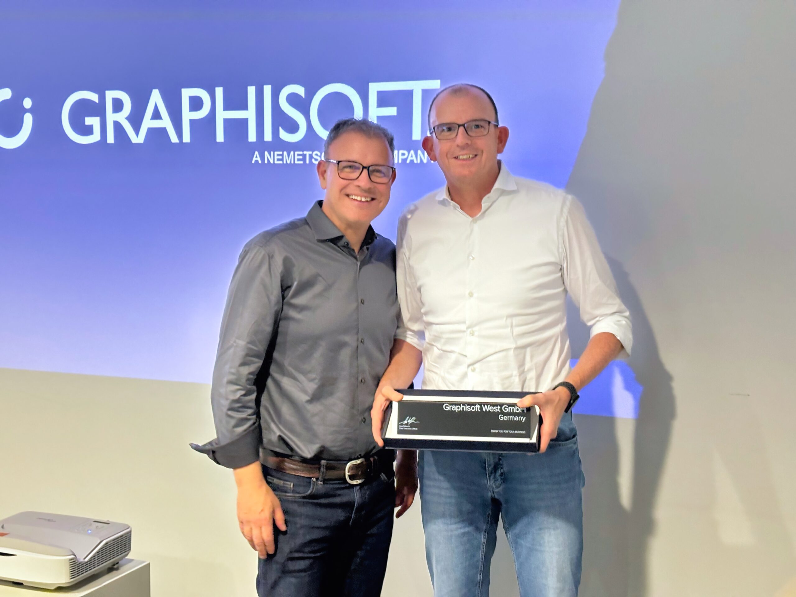 Zwei Personen stehen lächelnd nebeneinander und halten die SALES EXCELLENCE AWARD-Plakette. Im Hintergrund ist das Graphisoft-Logo im GRAPHISOFT CENTER WEST deutlich zu sehen.